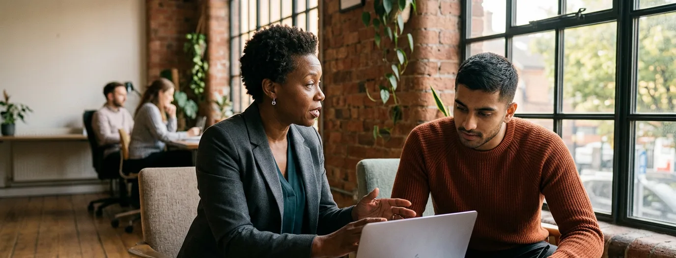 A mentor talking a younger professional through a plan on a laptop, in a brick loft with tall windows